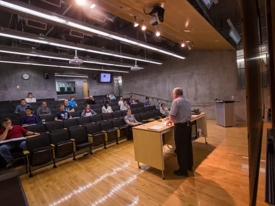 A man giving a lecture in a lecture hall, surrounded by students and using a microphone.