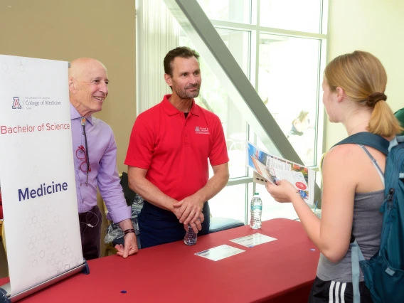 Two individuals stand beside a table displaying a sign, engaged in conversation or discussion.