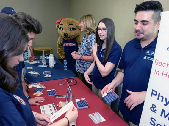 Several individuals are gathered around a table, enjoying the presence of a mascot, fostering a sense of community and fun.