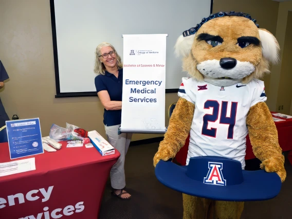 A woman poses beside a mascot at a table, smiling and engaging with the audience in a lively setting.