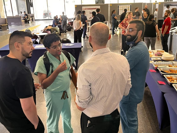 [A group of internal medicine residents, including Sanjeeva Weerasinghe, MD (PGY-3 – in light green scrubs), and Grant Collins, MD (PGY-2 – in white shirt), chat as food and beverages are served at the 2025 IM Research Academic Half-Day competition.]
