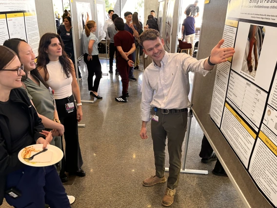 [Nathan Walton, MD, a PGY-2, talks about his poster to three IM residents, Jessica Kitsen, DO (left), Jackie Maltagliati, DO (center left), and Shenar Dinkha, DO (on right at left), all PGY-1s.]