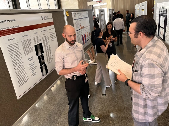[Division of Infectious Diseases’ Alex Perry, MD, MPH, and poster author Grant Collins, MD, a PGY-2 IM resident, discuss his poster, “1 View is no View,” about a 17-year-old male injured while playing football and the need for physical exams as well as imaging in such cases.]
