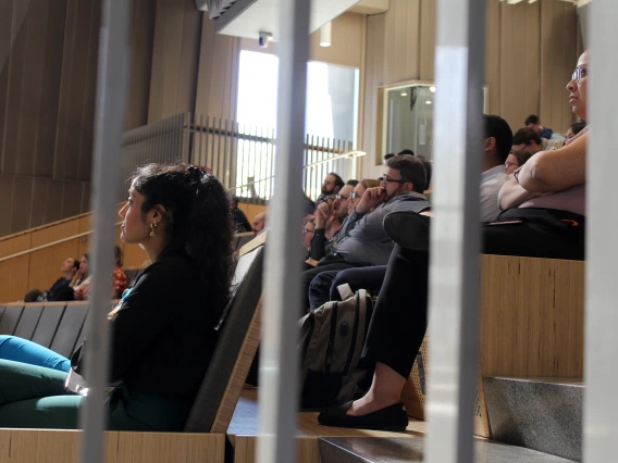 [PGY-2 IM resident Amrutha Doniparthi, MD (left) listens to the final Oral Presentation, as the judges (one row behind) consider the merits of the presenter’s research and presentation. She was among six residents who won for Best QI Project: “Improving pneumococcal vaccination rates in the resident clinic.”]