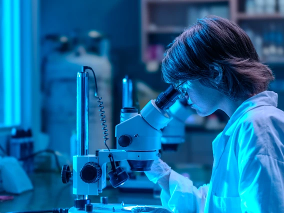 a young student is wearing a white lab coat and looking into a microscope. The image is tinted blue