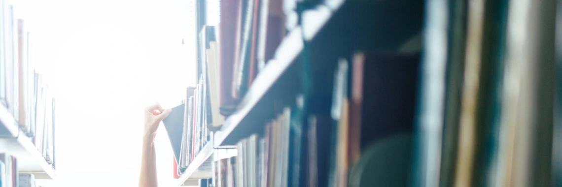 A person stretching their arm to reach for a book on a high shelf in a library setting.