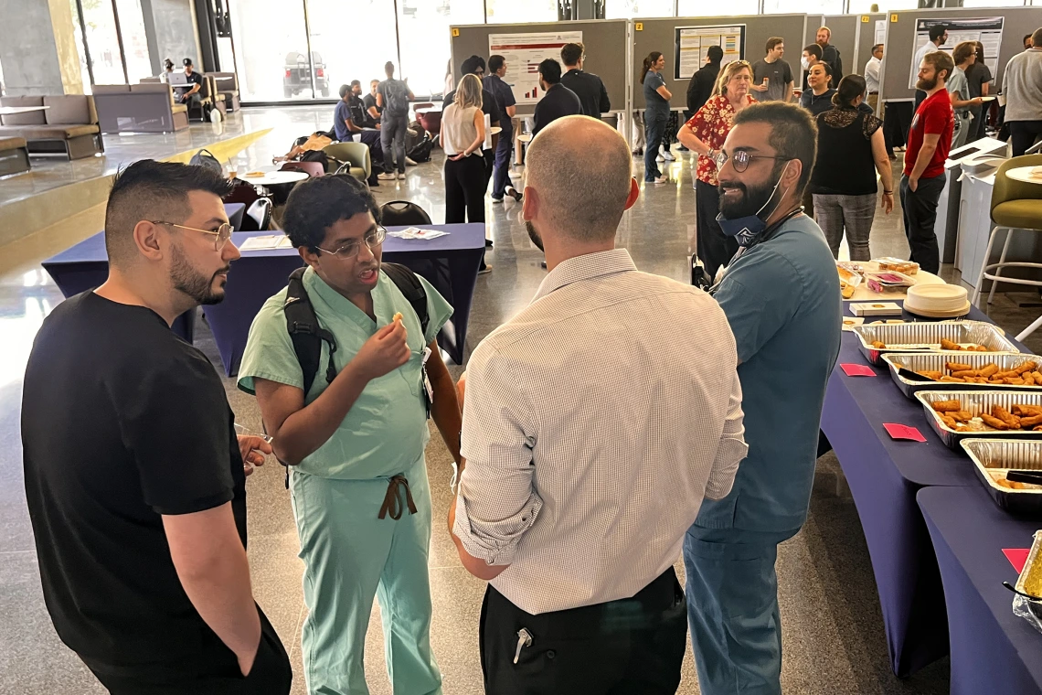[A group of internal medicine residents, including Sanjeeva Weerasinghe, MD (PGY-3 – in light green scrubs), and Grant Collins, MD (PGY-2 – in white shirt), chat as food and beverages are served at the 2025 IM Research Academic Half-Day competition.]