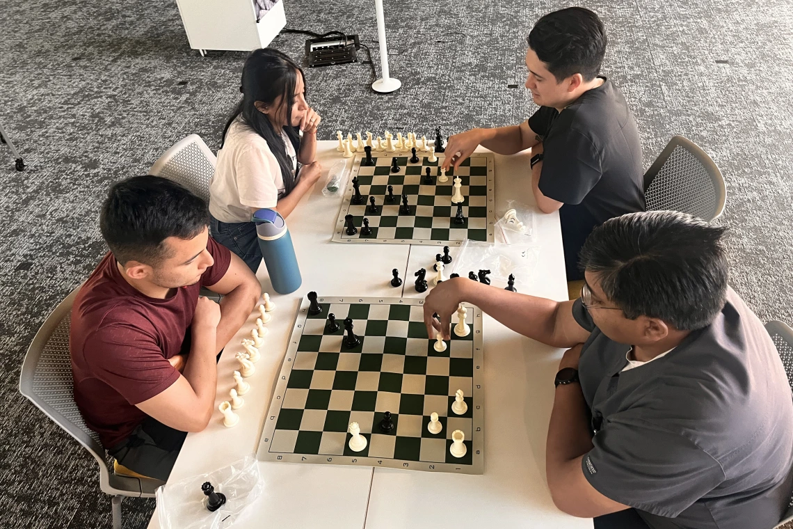 [Nathan Holt, DO (in black top – PGY-1 radiology resident), and Inpatient Medicine’s Anil Potharaju, MD (to Dr. Holt’s left), both decide to move their pawns at same time. The question is to which square.]
