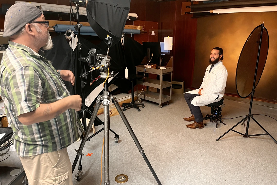 [UAHS BioCommunications photographer Mitch Masilun (left) prepares to shoot a white coat profile shot of Ethan Renfrow, DO, in the BioCom studio on the college’s third floor.]