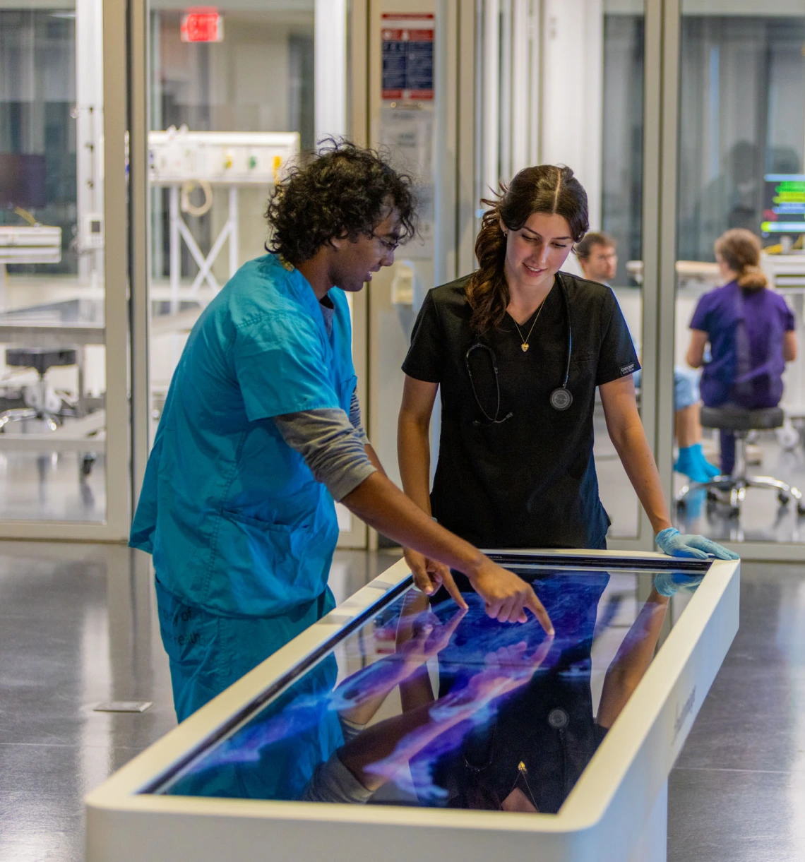 Two students in medical scrubs standing over a technology display of a human anatomy model