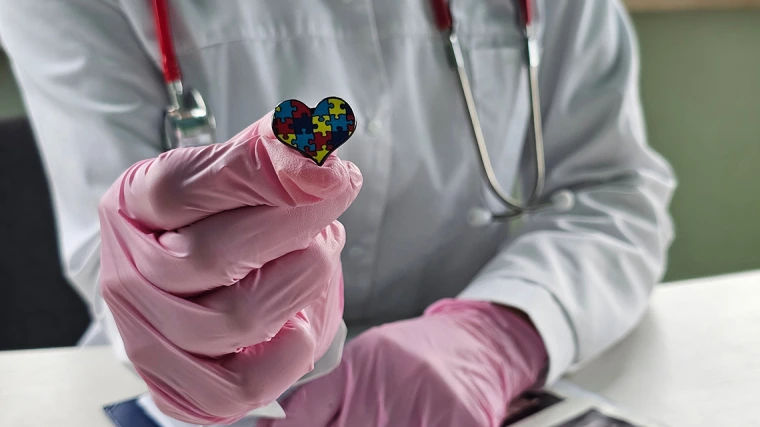 [An image, from the shoulders down, of a physician wearing a white coat, stethoscope and pink latex gloves while holding a small heart with a multi-color jigsaw puzzle design. (Courtesy: Adobe Stock)]