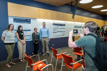 [Jennifer Stern, PhD, with students who work in her Endocrinology lab — Isabella Byington, Kassandra Bruner and Thadeus Carolyn — get a photo taken of them in front of one of their research posters at the SOAR in DOM forum.  ]